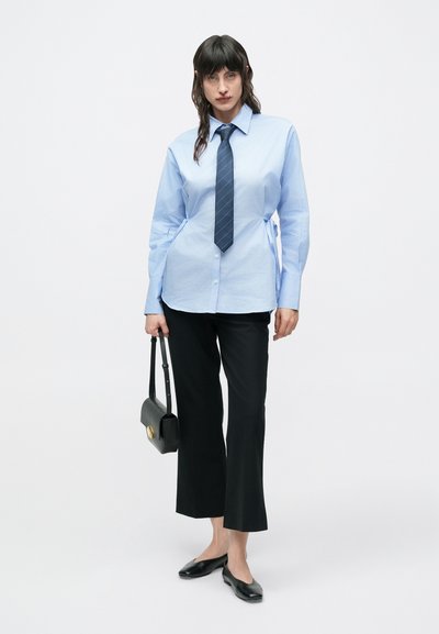 Ανοιχτό μπλε πουκάμισο με γιακά και μακριά μανίκια, σε συνδυασμό με μαύρο καμπάνα παντελόνι, μαύρες μπαλαρίνες και μικρή μαύρη τσάντα χειρός.