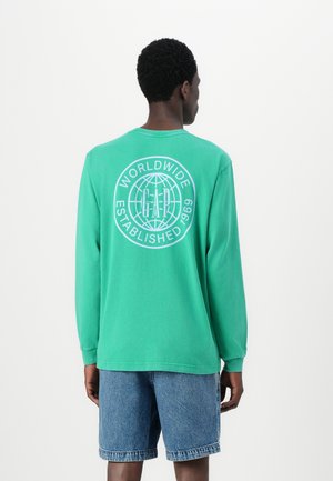Long-sleeve, green cotton t-shirt featuring a white globe graphic with "GAP" text on the back, paired with blue denim shorts.