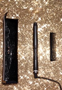 Un outil de coiffure noir avec un barillet effilé, un bouton marche/arrêt, un boîtier noir texturé et un peigne noir, le tout posé sur un fond doré pailleté.