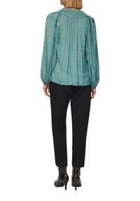 Türkise Plissierte Bluse mit langen Ärmeln und gerafftem Ausschnitt, kombiniert mit schwarzen schmalen Hosen und schwarzen High-Heel-Stiefeln.