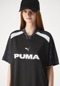 Camiseta desportiva preta de mangas curtas com decote em V, apresentando detalhes brancos, um grande logótipo "PUMA" e um padrão texturizado nas mangas. Boné preto incluído.