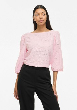 Kvinde iført en lyserød bluse med hulmønster og puffede 3/4-ærmer samt sorte bukser med høj talje, stående foran en ensartet hvid baggrund.