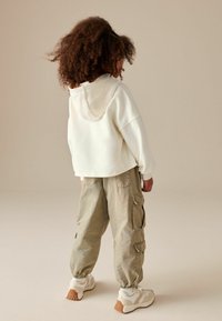 Weißer, kürzer geschnittener Hoodie mit Kapuze, kombiniert mit grünen Cargo-Hosen mit Seitentaschen und elastischen Bündchen. Beige Sneaker runden den Look ab.
