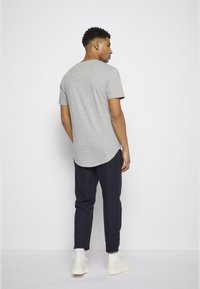 T-shirt cinza de manga curta com bainha curva, combinado com calças taper navy e ténis brancos. Modelo visto de costas.