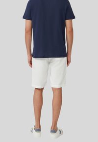 Marineblaues Baumwoll-T-Shirt kombiniert mit weißen Leinen-Shorts mit umgeschlagenen Ärmeln. Die Schuhe sind weiße Sneakers mit grauen Akzenten.