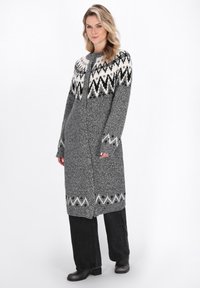 Lange grijze gebreide cardigan met een wit en zwart geometrisch patroon op de schouders, met een rechte snit en zijzakken. Zwarte broek en laarzen.