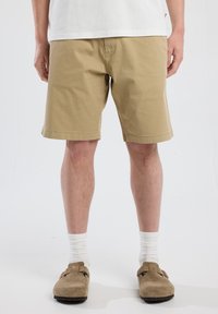 Beige cotton shorts with side pockets, mid-thigh length, and a straight fit. Worn with white socks and brown suede clogs.