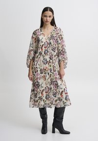 Florales Midikleid mit V-Ausschnitt, langen Puffärmeln, gefalteter Textur, auf einem cremefarbenen Hintergrund mit rosa, lila und roten Mustern. Schwarze kniehohe Stiefel.