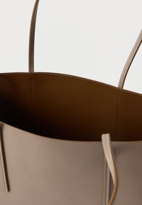 Beige bőrtáska barna belső béléssel és vékony vállpántokkal, felülnézetből bemutatva, amely felfedi a nyitott belső terét.