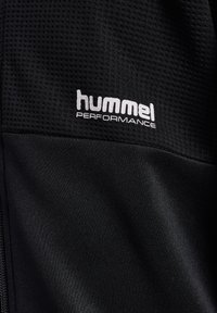 Czarna, sportowa tkanina z teksturowanym siatkowym obszarem. Posiada białe logo "hummel PERFORMANCE" znajdujące się po prawej stronie. Gładki, lekki materiał.