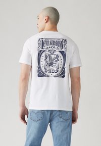 Tricou alb cu maneci scurte, având un grafic albastru pe spate cu un design de vultur și textul "Levi Strauss & Co.". Material din bumbac moale.