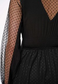 Zwarte jurk met transparante, stipjesmouwen en een smocktail. De stof heeft een zachte textuur met een gelaagde zwarte tule rok.