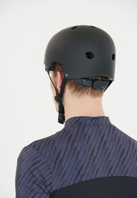 Svart hjälm med ventilationshål, justerbar rem och bakhjul. Bärs över en mönstrad mörkblå skjorta med vertikala ränder.