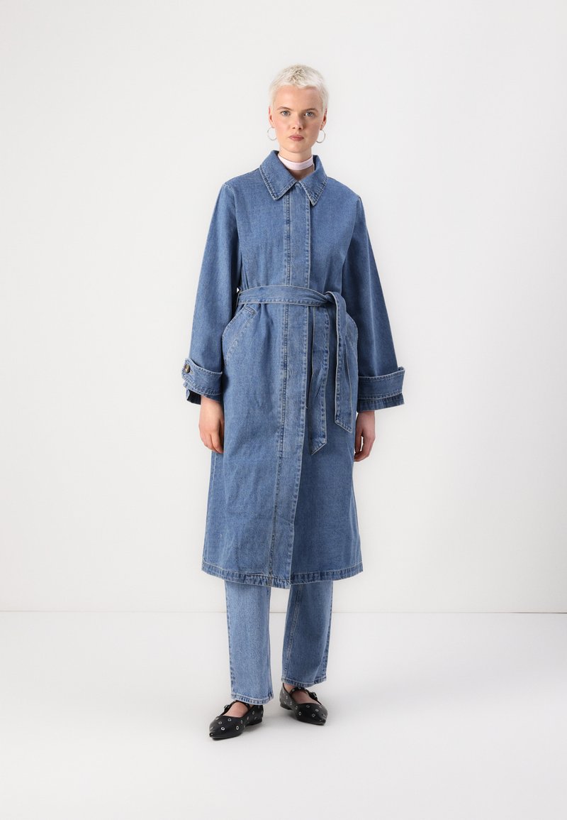 Cappotto di jeans con maniche lunghe, cintura in vita, due tasche laterali e colletto. Colore azzurro chiaro, orlo leggermente sfilacciato.
