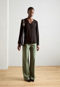 Camicia nera a maniche lunghe con scollatura a V, abbinata a pantaloni larghi verdi e scarpe a punta nere. Il modello porta una borsa nera con hardware dorato.