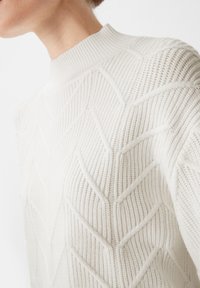 Cremfarbener Pullover mit hohem Kragen. Der Stoff weist ein strukturiertes Blattmuster und gerippte Details auf, die ein strukturiertes Design betonen.