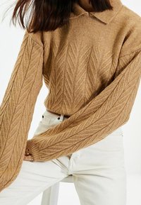 Suéter de punto marrón con un cuello con solapa, que presenta un patrón de espiga texturizado y mangas anchas, combinado con pantalones blancos de cintura alta.