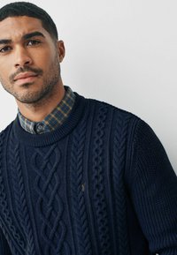 Marineblå kabelstrikket sweater med rund hals, prydet med flettede mønstre og en lille logo-detalje. Laget over en ternet skjorte.
