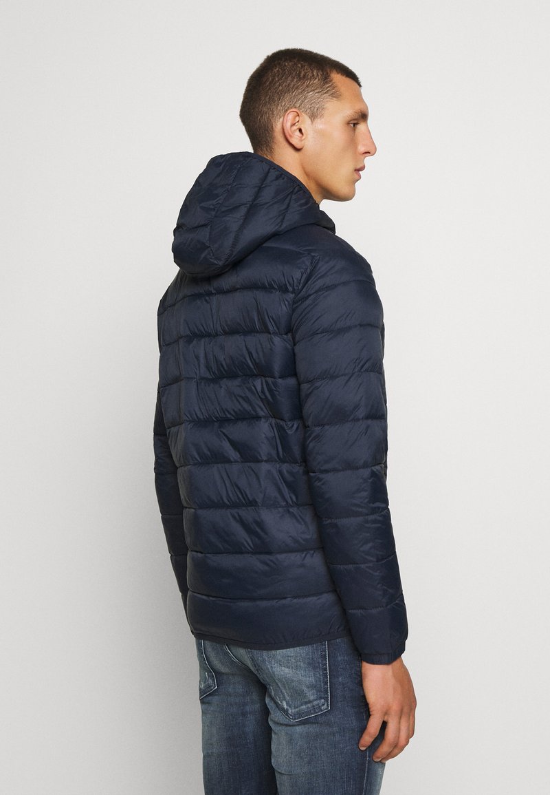 Veste matelassée marine avec capuche, présentant un design en quilt, une texture lisse et des poignets élastiques. Associée à un jean bleu pour un look décontracté.