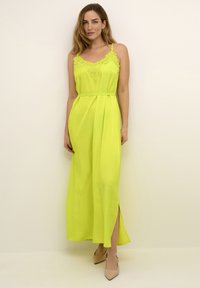 Femeie îmbrăcată într-o rochie maxi fără mâneci, de culoare verde lime, cu detalii din dantelă, pe un fundal neutru, asortată cu pantofi cu vârf ascuțit de culoare bej.