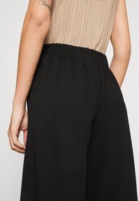 Pantalon noir à jambes larges avec une taille élastique, en tissu lisse et texturé. Gros plan sur l'arrière et une main près de la poche.