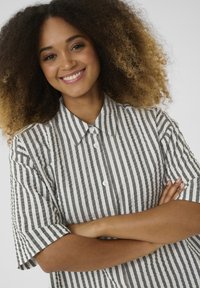 Camicetta a maniche corte a righe grigia e bianca, con collo a camicia, dettagli con bottoni e una vestibilità rilassata. Tessuto testurizzato visibile.