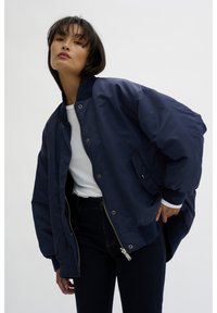 Navy blue bomber jacket with a shiny finish, zip closure, ribbed cuffs, and a side pocket. Worn over a white top and dark jeans.