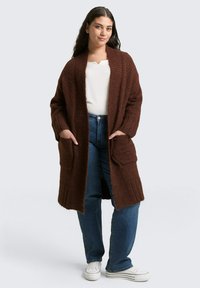 Brązowy dzianinowy kardigan o luźnym kroju, z długimi rękawami, otwartym przodem i dwoma przednimi kieszeniami, noszony na białej bluzce i niebieskich dżinsach.