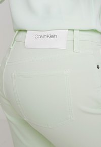 Acercamiento al bolsillo trasero de unos vaqueros verde claro con etiqueta de cuero blanca de la marca Calvin Klein en la cinturilla.