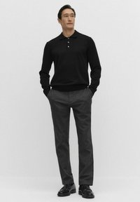 Czarna koszula polo z długim rękawem i trzema guzikami, w połączeniu z ciemnoszarymi spodniami. Mężczyzna stoi z rękami w kieszeniach, nosząc czarne buty.