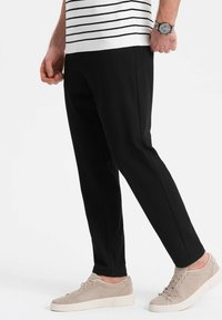 Zwarte taps toelopende broek met zijnaad, gecombineerd met lichtbeige suède sneakers met veters en een gestructureerde afwerking. Gestreept shirt zichtbaar bovenop.