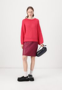 Rosa gestrickter Pullover mit langen Ärmeln, kombiniert mit einem figurbetonten burgunderfarbenen Lederröcke. Das Model trägt schwarze Plateau-Clogs und hält eine schwarze Handtasche.