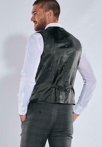 Schwarze Satinweste mit tailliertem Schnitt und einer Schnalle am Rücken. Getragen über einem weißen Hemd, kombiniert mit grau karierten Hosen.