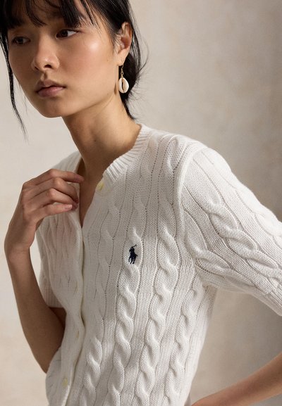 Młoda kobieta o ciemnych włosach nosi biały krótki kardigan w warkocze i kolczyk w kształcie muszli, stojąc na neutralnym tle.