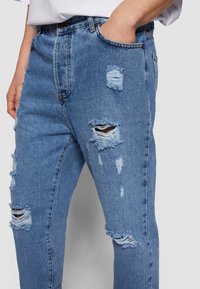 Jeans en denim bleu avec un design déchiré et plusieurs déchirures effilochées. Le tissu a un lavage moyen et une texture légèrement décolorée.