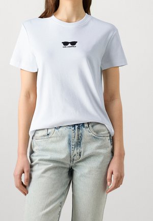 Kvinde iført hvid Karl Lagerfeld t-shirt med sort solbrillelogo og lyse vaskede jeans, stående mod en ensfarvet baggrund.