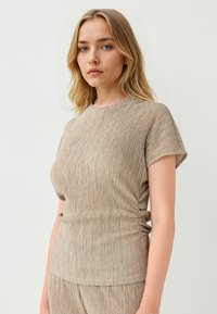 Beige geribbelde gebreide top met een ronde halslijn en korte mouwen. Heeft zijkant rimpeling voor een getailleerde uitstraling. Zachte textuur en flexibel materiaal.