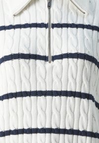 Jersey de color crema con cuello con cremallera; presenta patrones de trenzado texturizado y rayas horizontales en azul marino. Tejido suave.