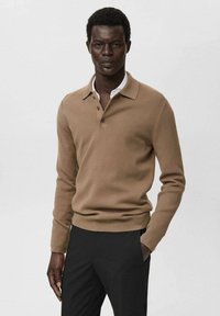 Male model wearing a camel-colored long-sleeve polo sweater and black trousers, standing with one hand in pocket against a plain background.