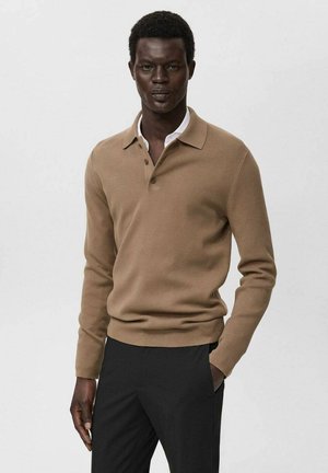 Modello maschile che indossa un maglione polo a maniche lunghe color cammello e pantaloni neri, in piedi con una mano in tasca su uno sfondo neutro.