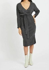 Šedé zavinovací šaty s dlouhými rukávy, výstřihem do V a zavázaným pasem. Výrazný postranní rozparek a strukturovaná pletenina. Nosené s bílými kotníkovými botami.