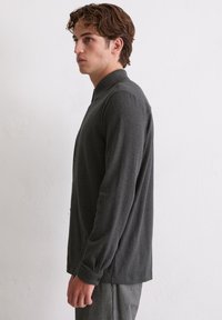 Młody mężczyzna o falistych brązowych włosach, ubrany w ciemnoszary koszulowy sweter z długim rękawem i szare spodnie, stojący bokiem przy białej ścianie.