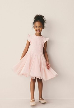 Young girl smiling while twirling in a light pink sleeveless dress with textured details and wearing matching ballet flats.