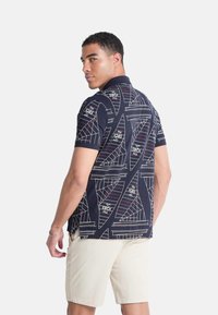 Polo de manga corta en tejido azul marino con patrones geométricos de veleros en blanco y rosa. El modelo lleva pantalones cortos beige.