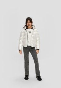Weiße, dicke, isolierte Jacke mit Kapuze; schwarzer Reißverschluss und Akzente. Kombiniert mit einem grafischen weißen Sweatshirt und dunkelgrauen Jeans.