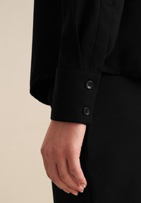 Zwart langarm shirt manchet met twee zwarte knopen, voorzien van een gladde stofstructuur en gestructureerd ontwerp. Hand rustend op het manchet.