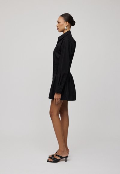 Abito camicia in cotone nero con bottoni frontali, maniche lunghe, vita elastica e gonna svasata. Indossato con sandali neri con tacco.