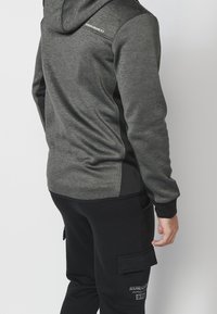 Sweat à capuche gris avec une finition texturée, coupe ajustée et panneaux latéraux noirs. Comprend un logo au dos et des poignets élastiques.