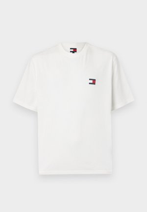 Valkoinen puuvillapaita, jossa on pyöreä kaula-aukko ja lyhyet hihat. Vasemmassa rinnassa on pieni logo-patch punaisena ja tummansinisena.