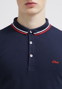 Ciemnoniebieska koszulka polo z dwoma guzikami, z kołnierzykiem w czerwono-białe paski i czerwoną haftowaną logo na piersi.